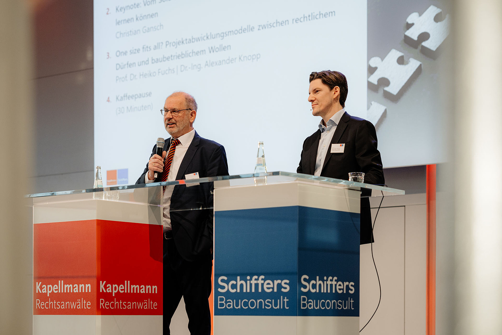 Schiffers-Bauconsult-Geschaeftsfuehrer-Alexander-Knopp-rechts-und-Anwalt-Werner-Langen-von-Kapellmann-Rechtsanwaelte-links-auf-der-Buehne-bei-einer-Podiumsdiskussion-waehrend-des-Kapellmann-Schiffers-Baukongress-2026